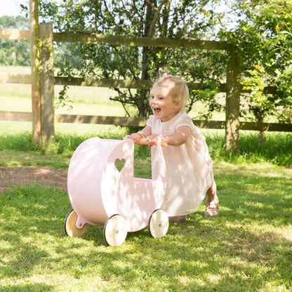 Smiling toddler in a cream dress pushing a pink wooden doll’s pram on the grass under dappled sunlight.