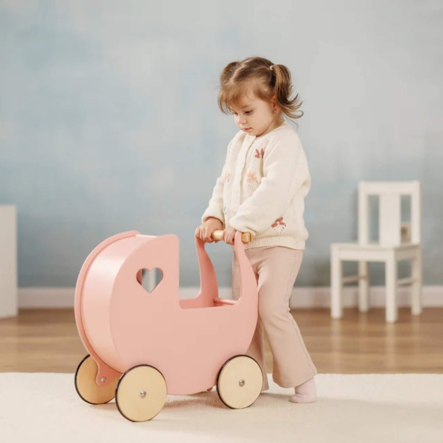 Young girl indoors holding the handle of a pink wooden doll’s pram, standing on a soft cream rug.