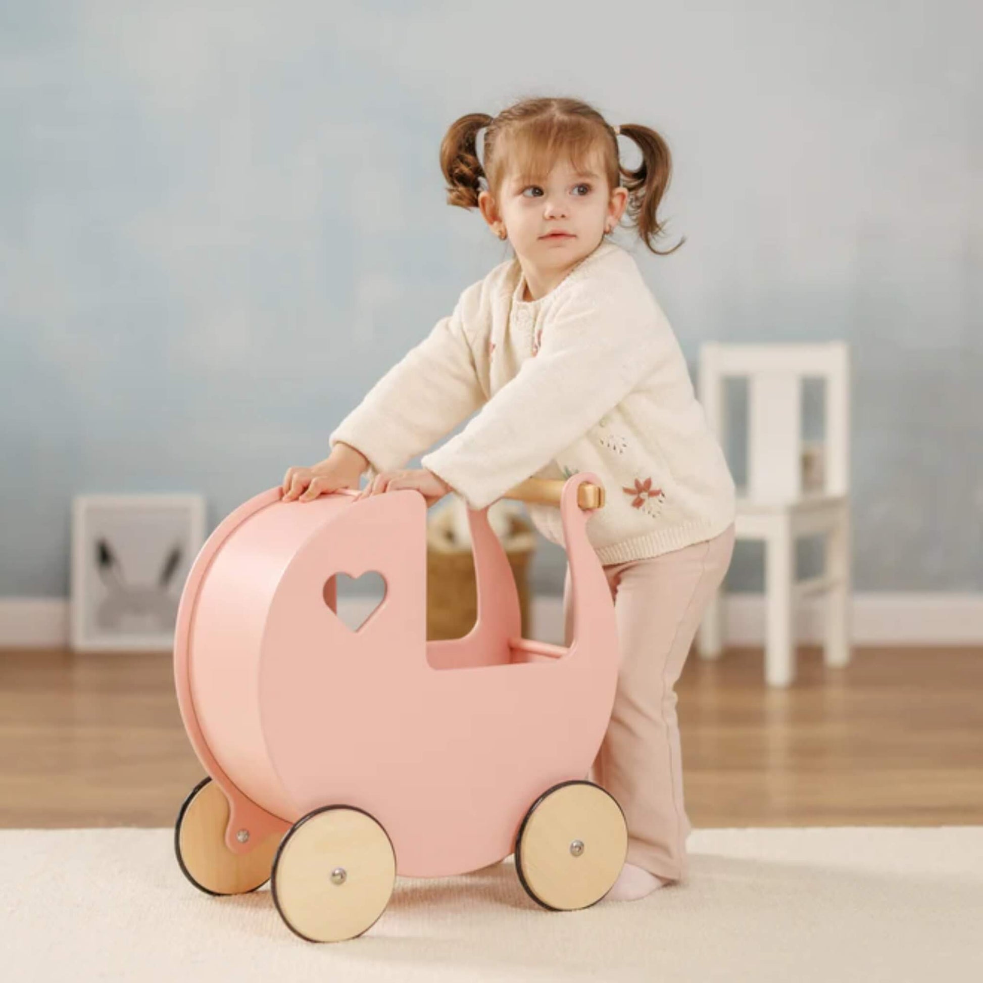 Little girl with pigtails pushing a pink wooden doll’s pram across a light wooden floor in a softly lit room.