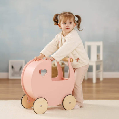 Little girl with pigtails pushing a pink wooden doll’s pram across a light wooden floor in a softly lit room.