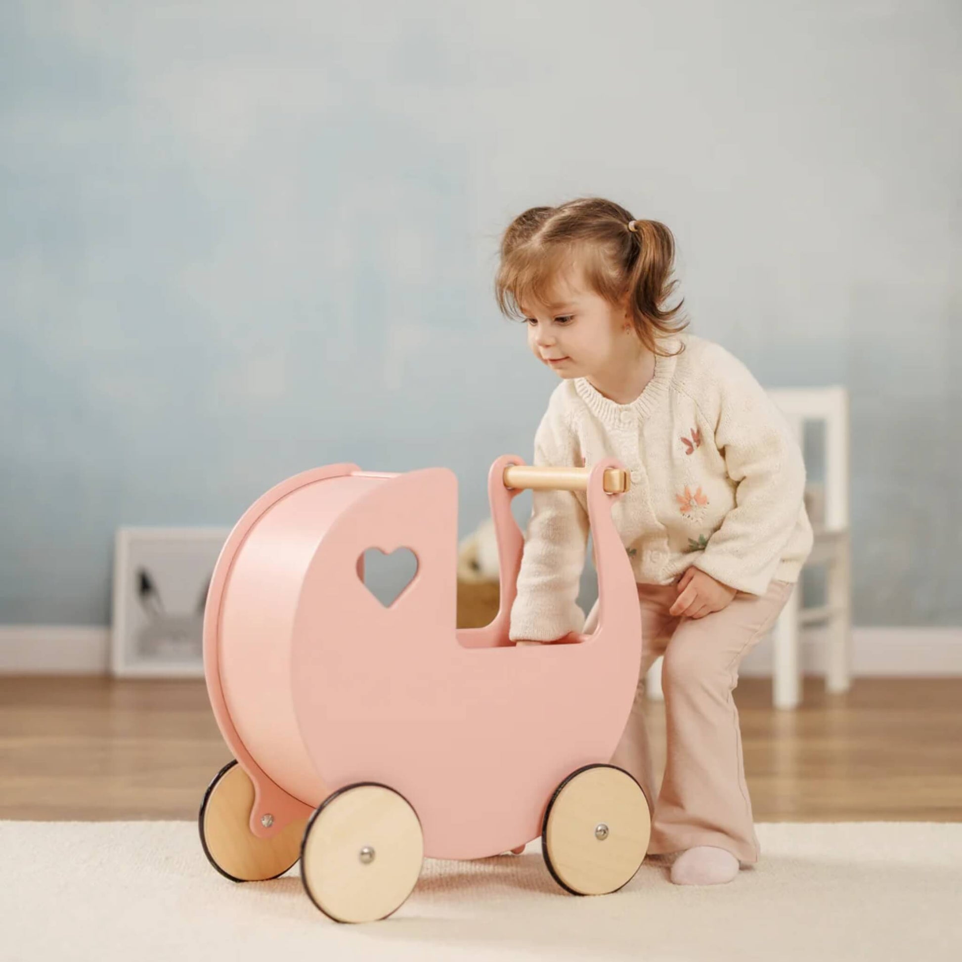 Child leaning forward to look inside a pink wooden doll’s pram in a softly decorated nursery room.