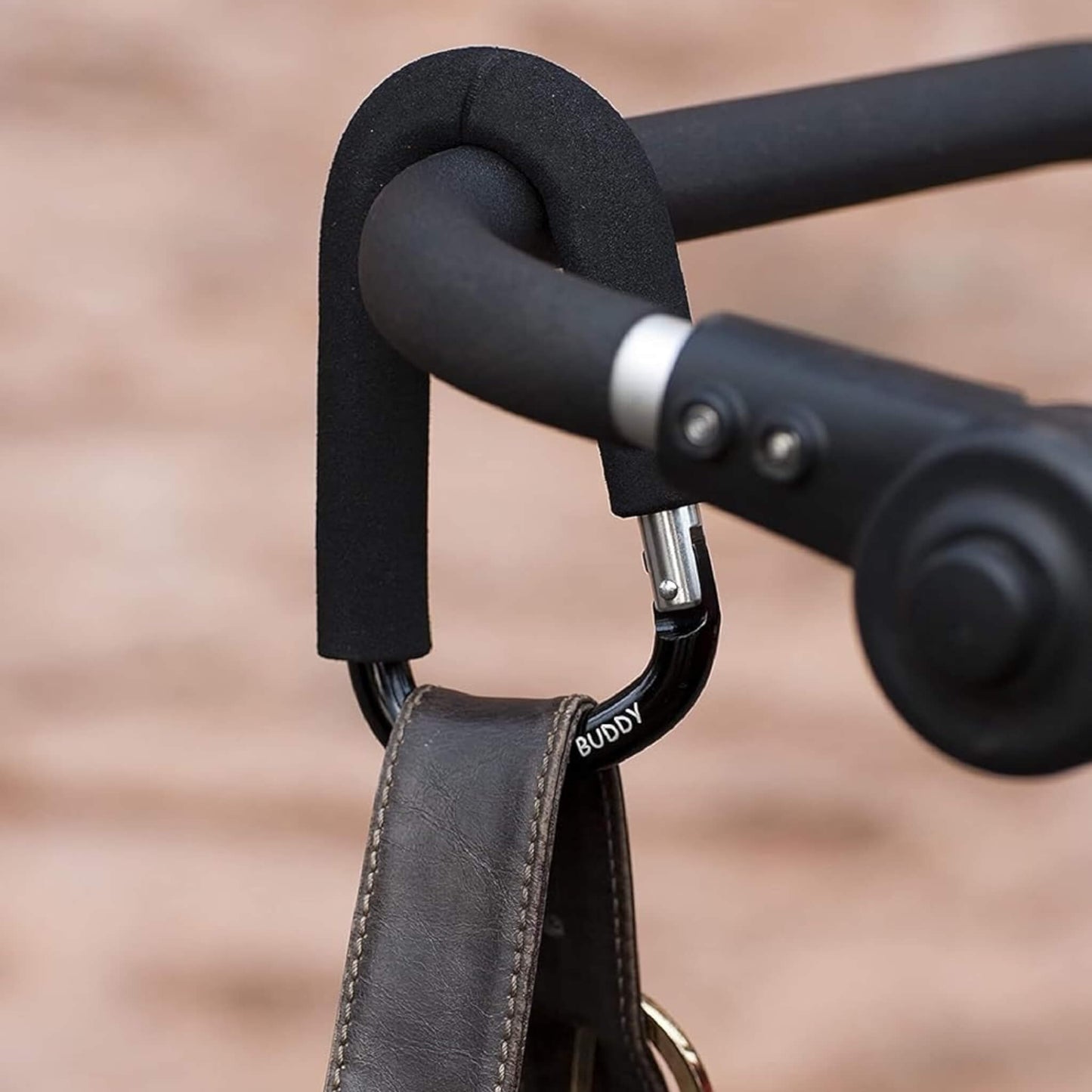 Close-up of foam-covered buggy clip fastened to a stroller handle, carrying a leather bag strap.