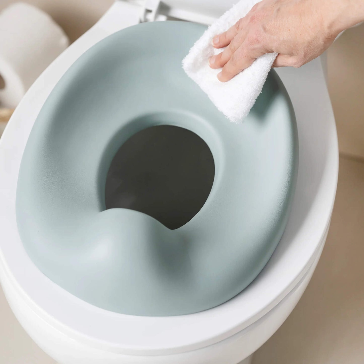 A hand wiping a soft green potty seat while it sits on a toilet, showing the smooth surface being cleaned.