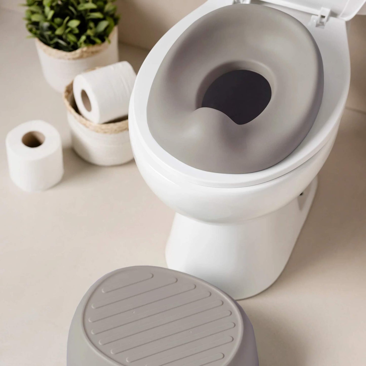 A taupe potty seat positioned on a standard toilet, with toilet paper rolls and small plant baskets in the background.