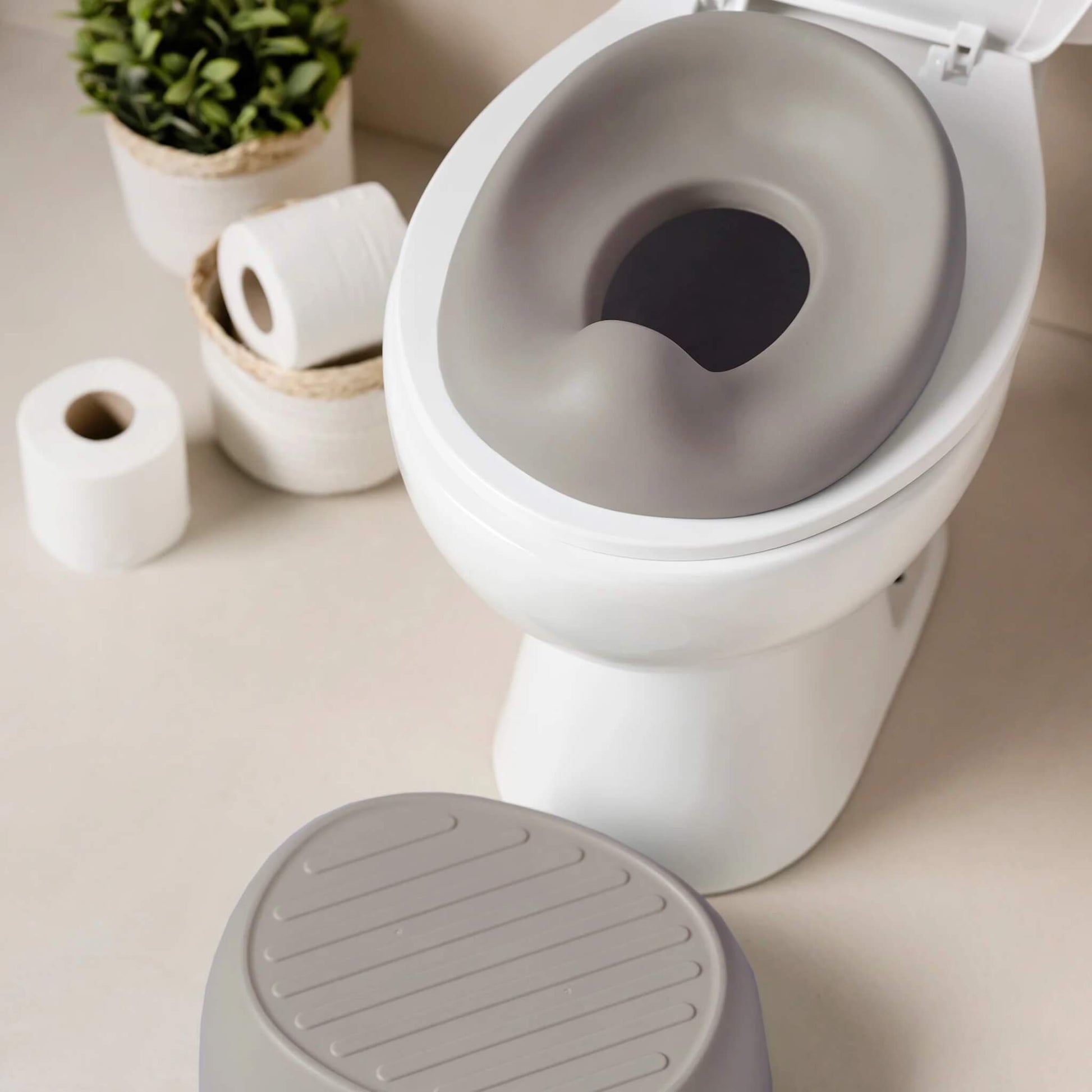 A taupe potty seat positioned on a standard toilet, with toilet paper rolls and small plant baskets in the background.