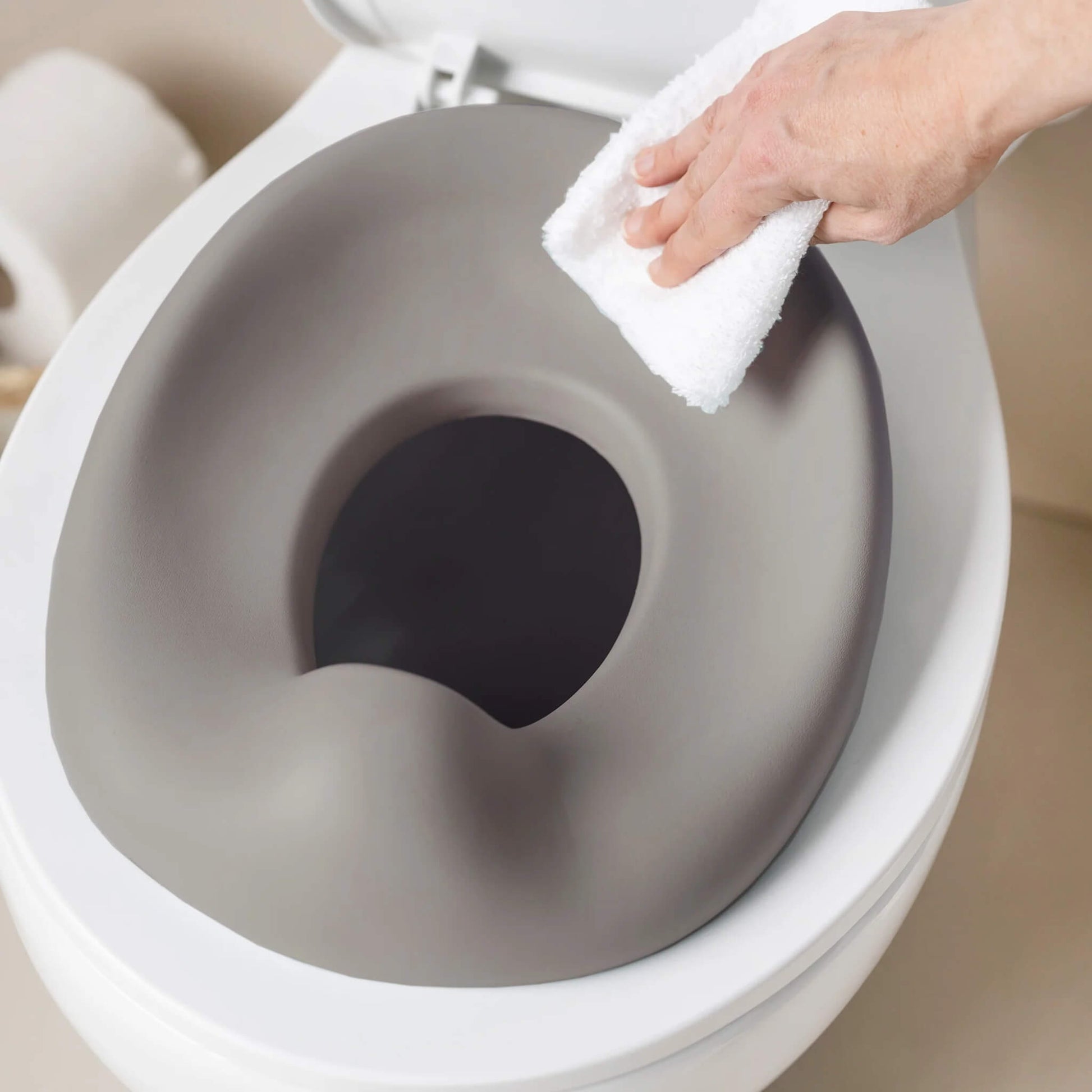 A hand wiping a taupe potty seat while it sits on a toilet, showing the smooth, easy-clean surface.