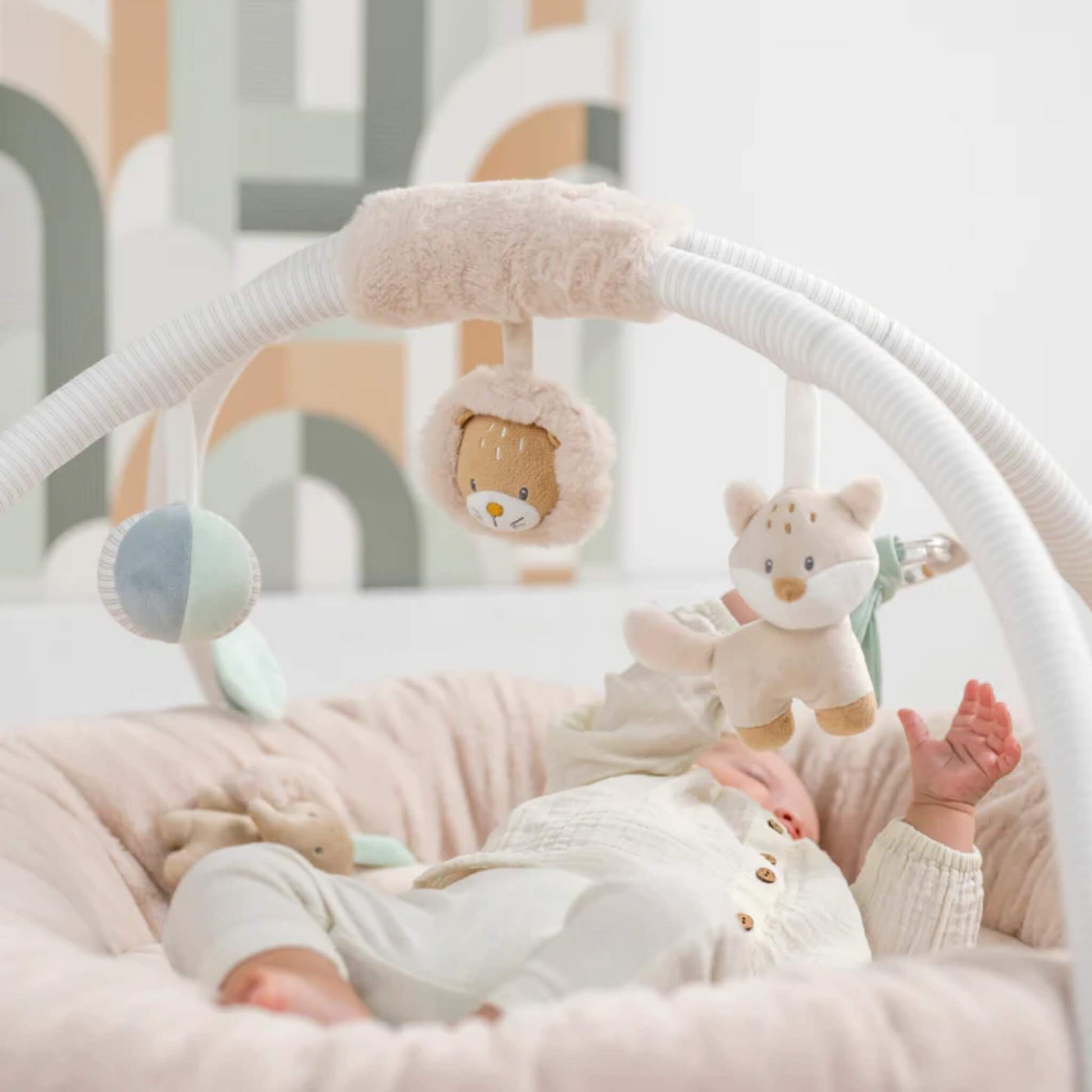 Baby lying comfortably inside a soft padded playmat reaching up toward hanging plush animal toys suspended from curved fabric arches.