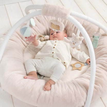 Baby stretching arms toward pastel fabric toys and mirror while lying inside a cushioned circular play gym with raised sides.