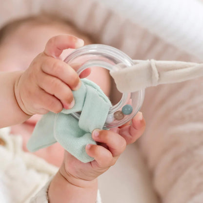 Small baby hand gripping a clear ring rattle toy wrapped with mint green fabric, showing textured sensory details for grasping and shaking.