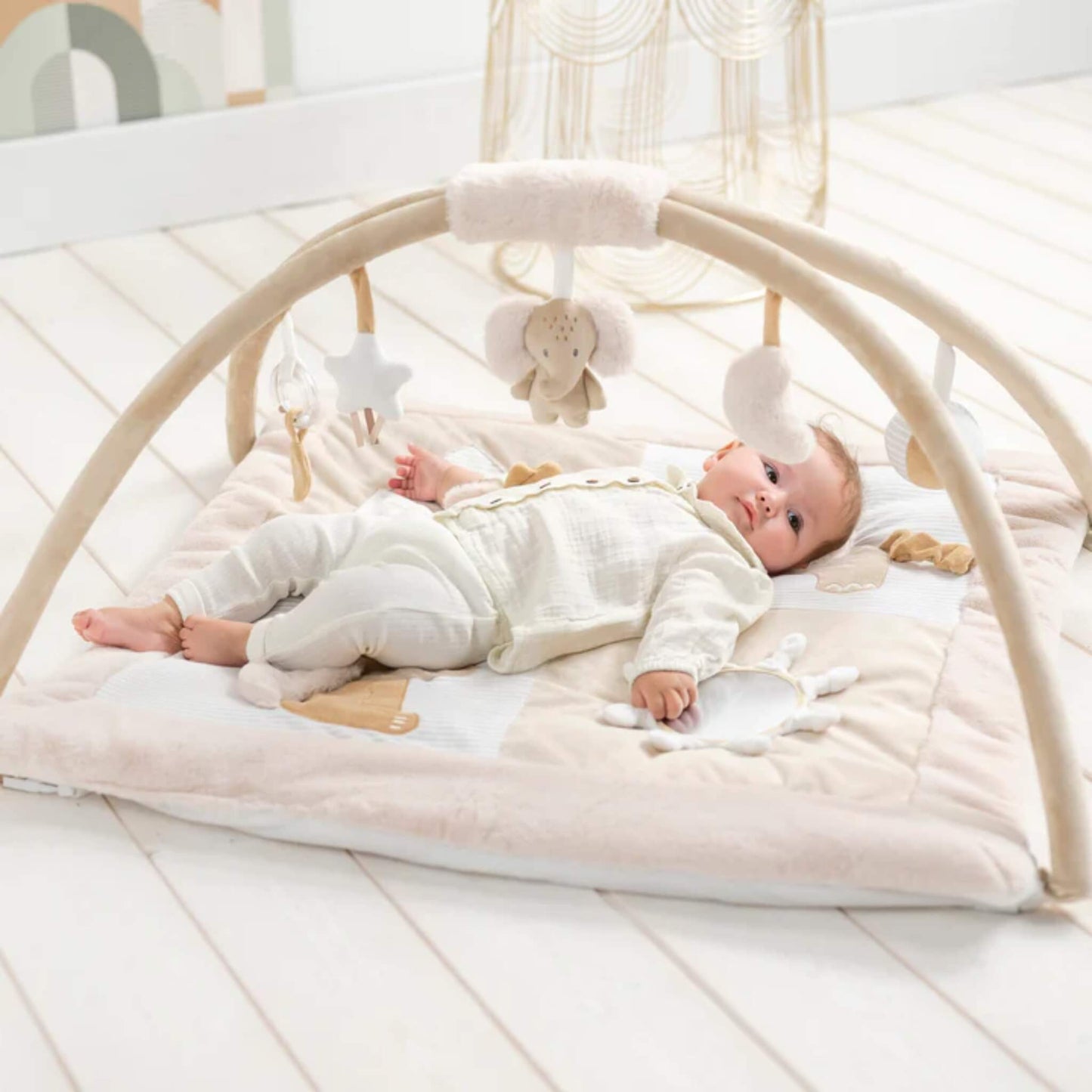 Baby lying relaxed on a padded fabric playmat beneath curved arches with hanging soft toys, set on a pale wooden nursery floor.