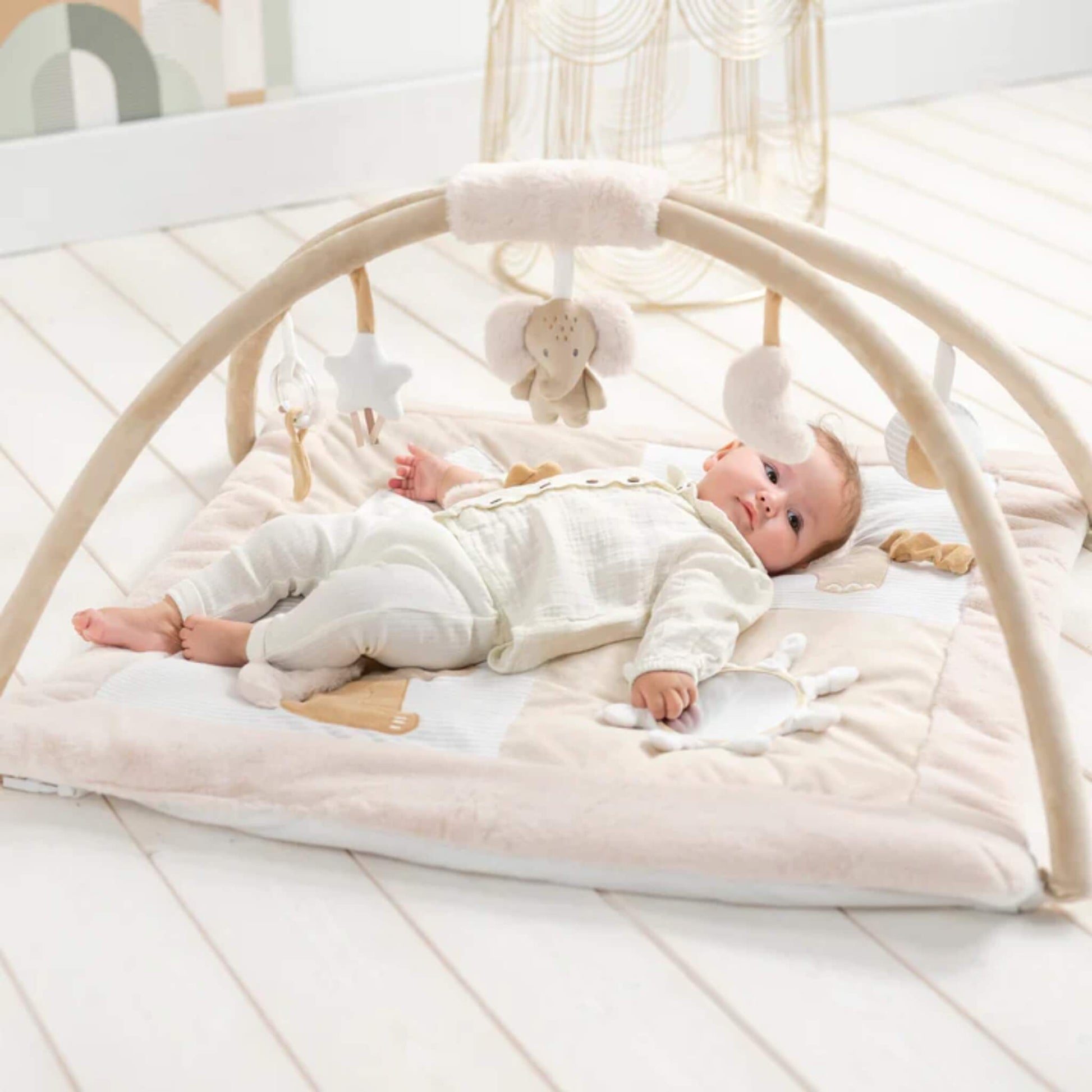 Baby lying relaxed on a padded fabric playmat beneath curved arches with hanging soft toys, set on a pale wooden nursery floor.