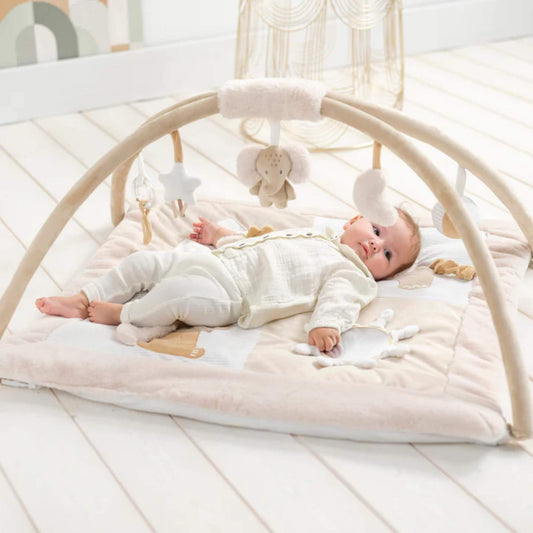 Baby lying relaxed on a padded fabric playmat beneath curved arches with hanging soft toys, set on a pale wooden nursery floor.