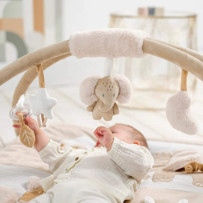 Baby lying on padded activity mat reaching up toward hanging soft toys suspended from curved fabric arches in a bright nursery setting.