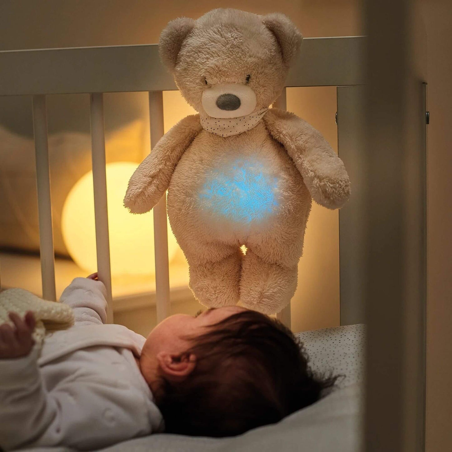 A plush cuddly bear nightlight hanging on a crib, glowing softly with a blue light as a baby lies in the cot below. 