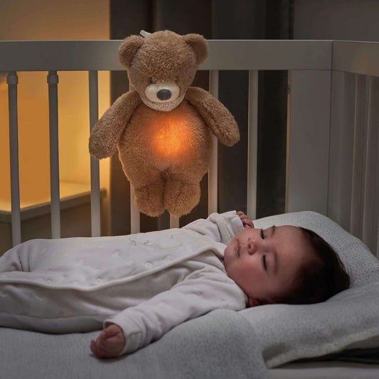 Plush soft bear nightlight hanging on a crib, glowing softly with an amber light as a baby lies in the cot below. 