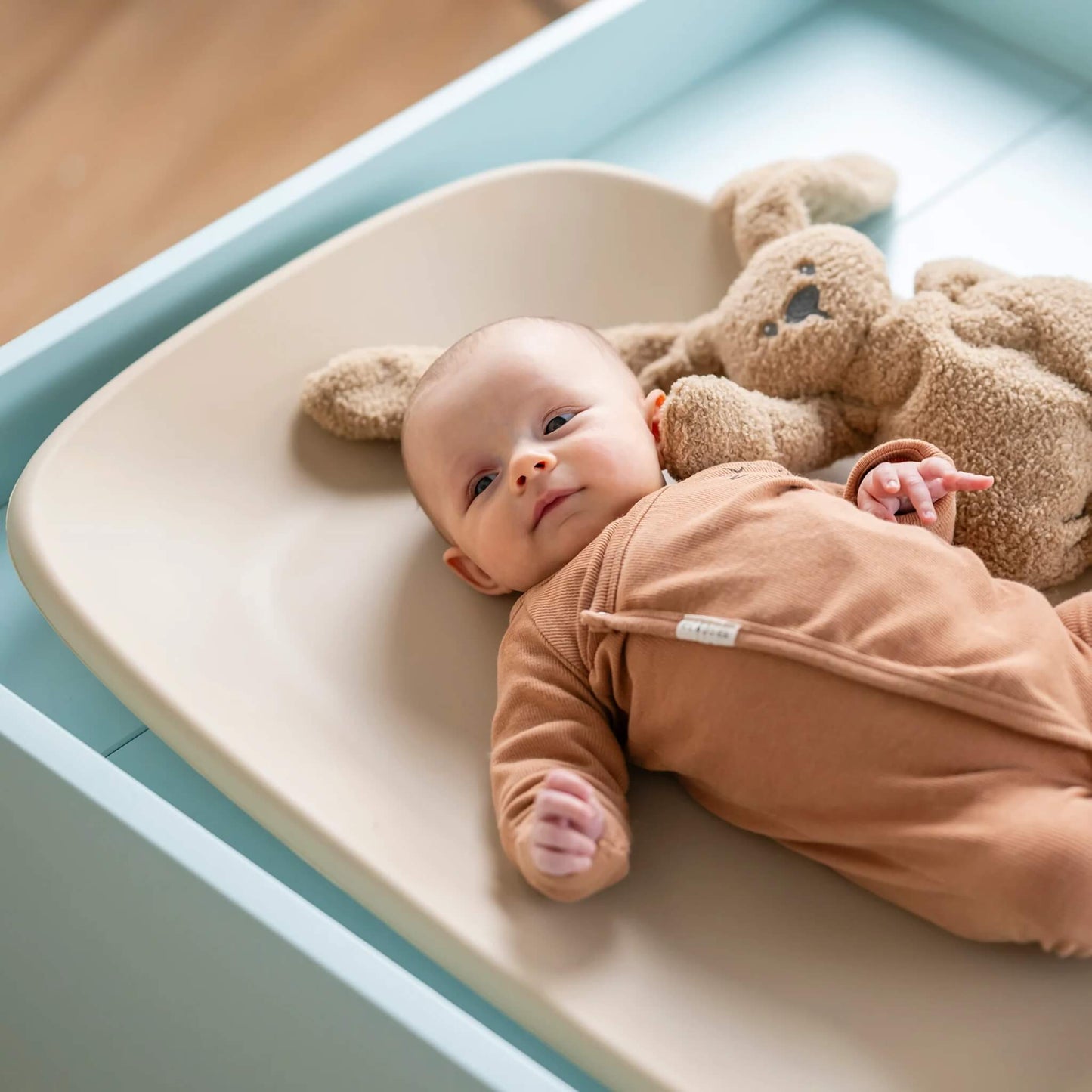 Baby lying on a beige contoured changing pad with a soft toy beside them, resting comfortably on the padded surface.