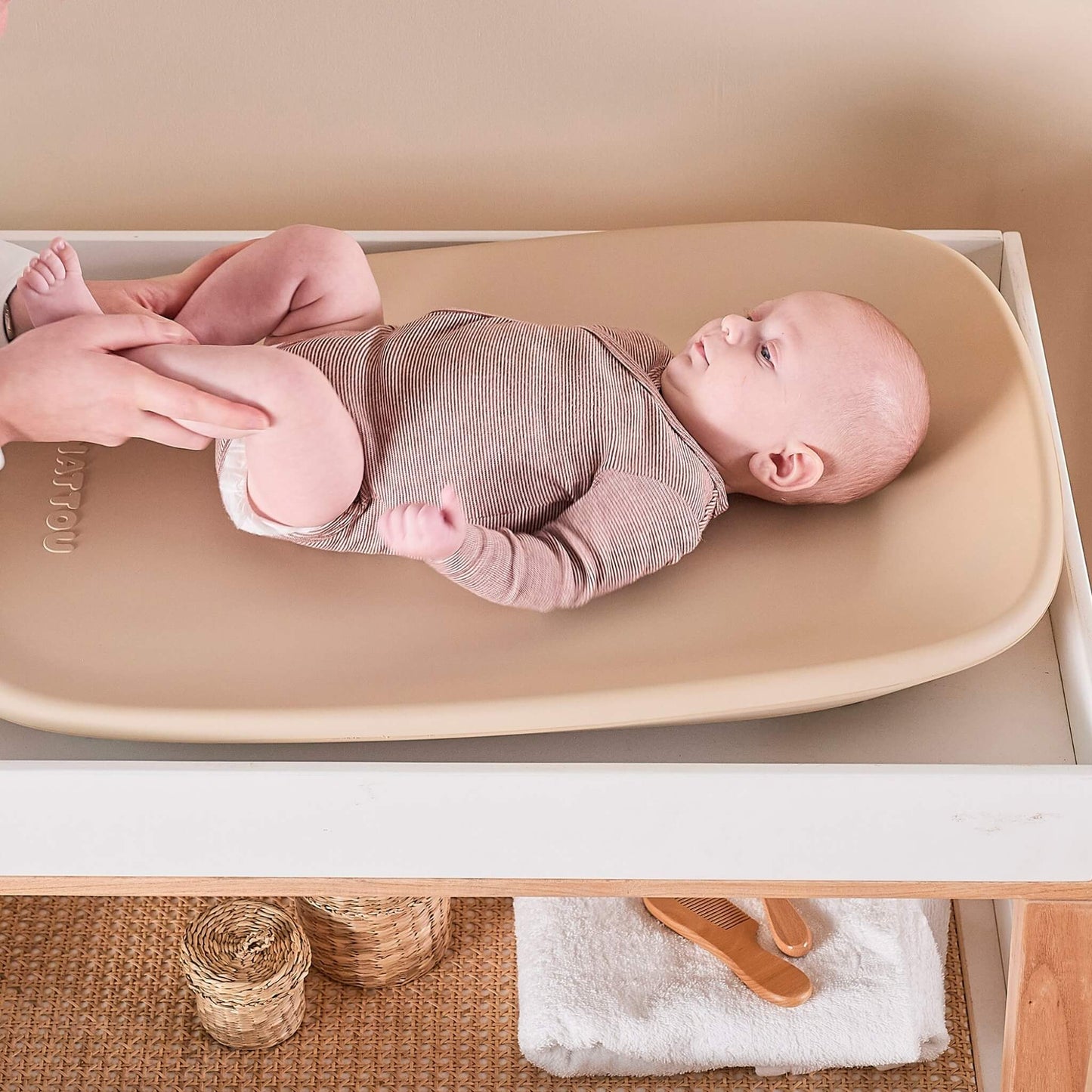 Baby lying on a beige contoured changing pad while an adult supports their legs during a nappy change.