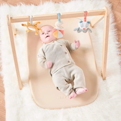 Baby lying on a beige changing pad placed beneath a wooden play gym with hanging toys above.