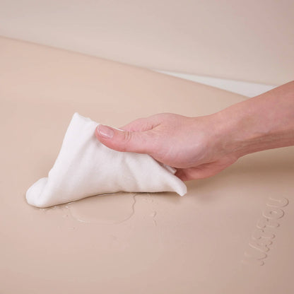 Adult hand using a soft cloth to wipe water from the surface of a beige changing pad.