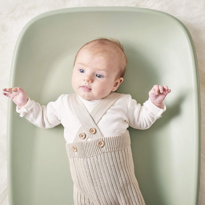 Baby lying on a soft green changing pad with arms relaxed and head supported by the curved surface.