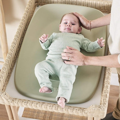 Baby lying on a soft green changing pad while an adult gently supports their head and torso during a nappy change.