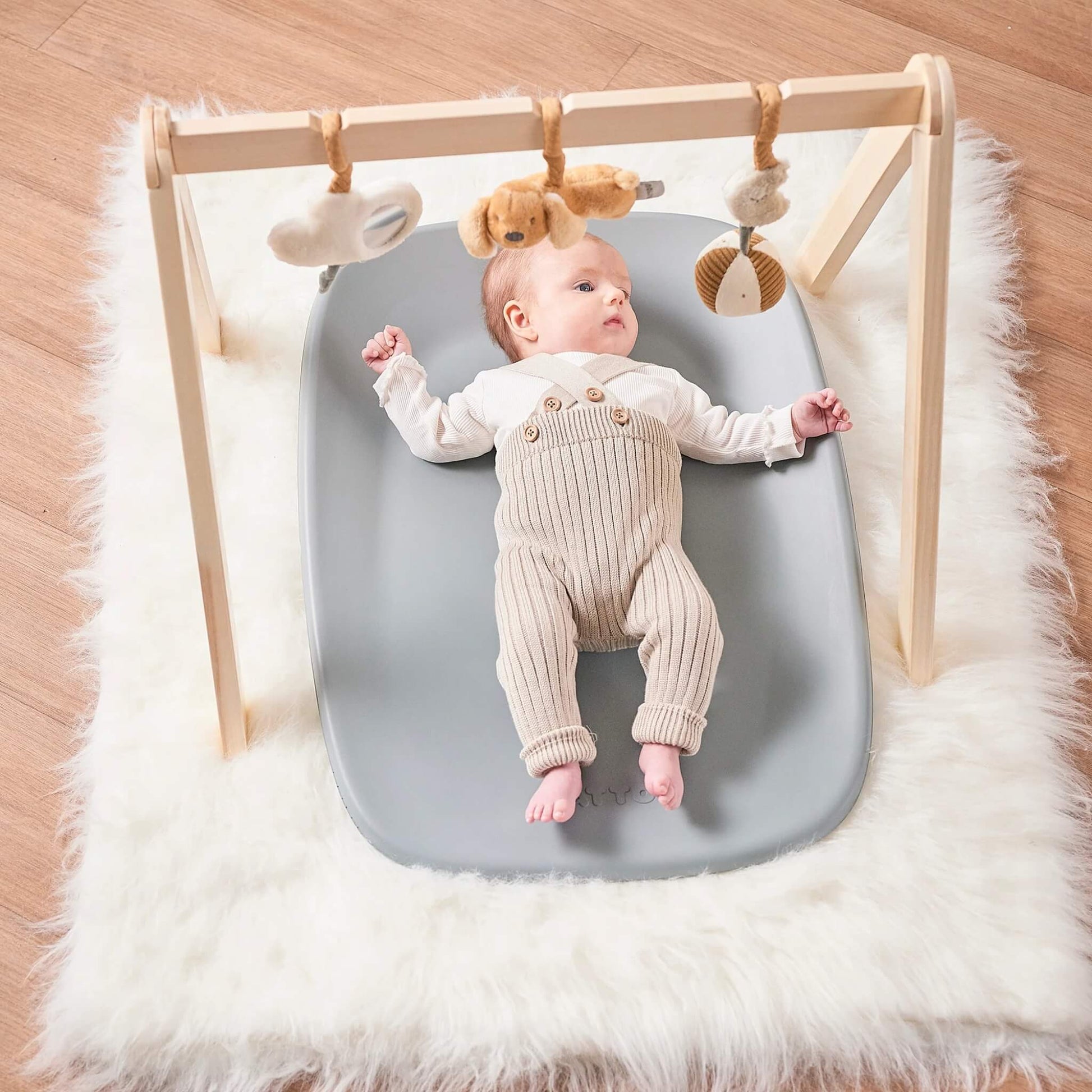 Baby lying on a grey changing pad beneath a wooden play gym with soft hanging toys.