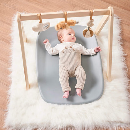 Baby lying on a grey changing pad beneath a wooden play gym with soft hanging toys.