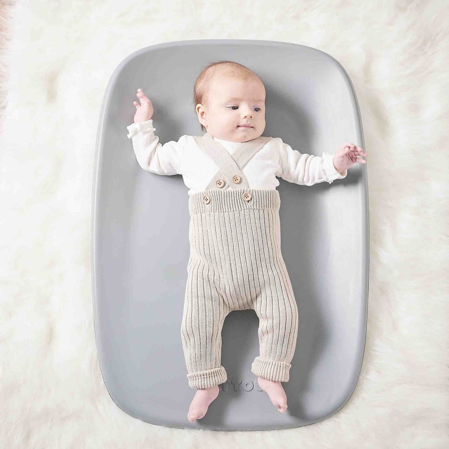 Baby lying on a grey changing pad placed on a soft, fluffy surface, arms and legs gently extended.