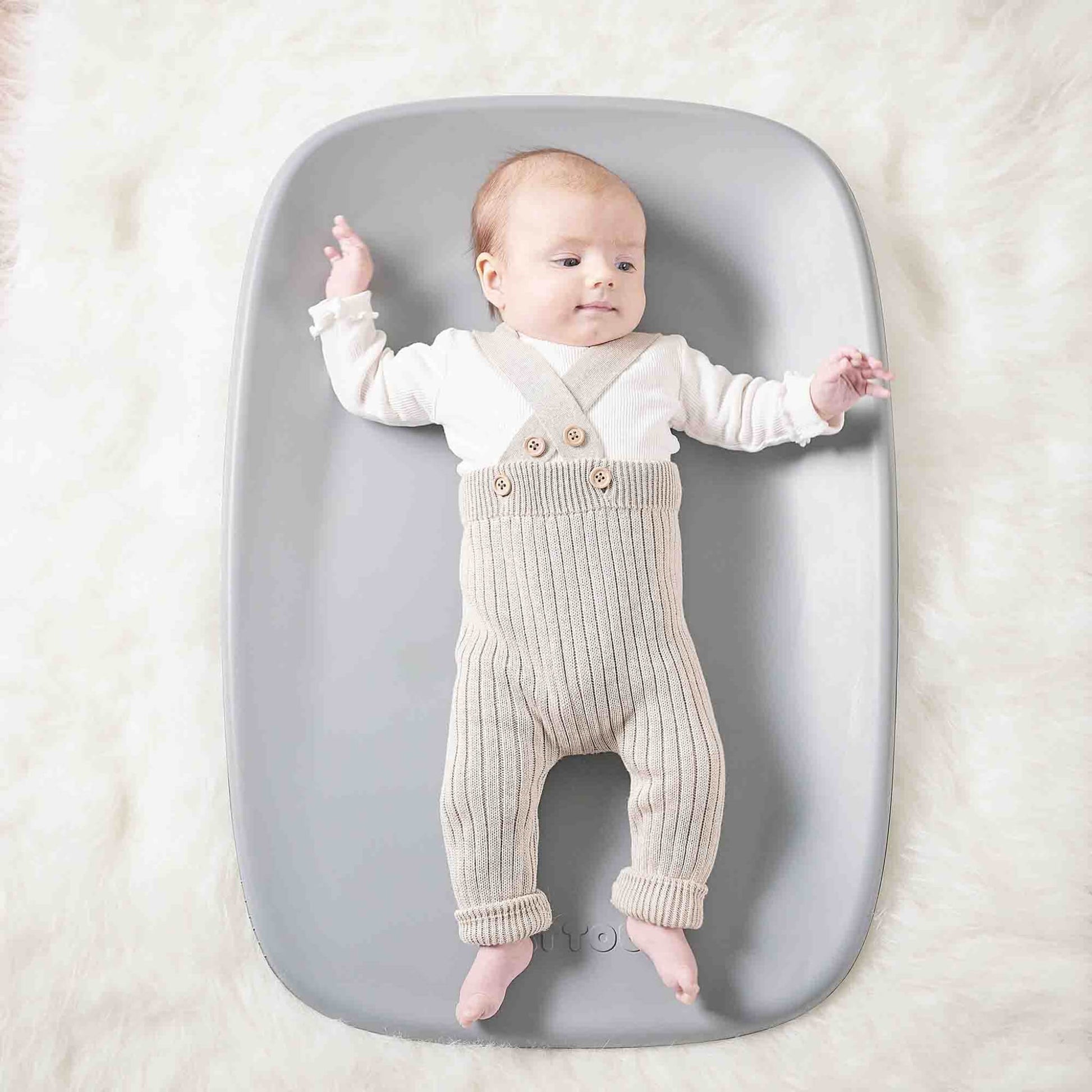 Baby lying on a grey changing pad placed on a soft, fluffy surface, arms and legs gently extended.