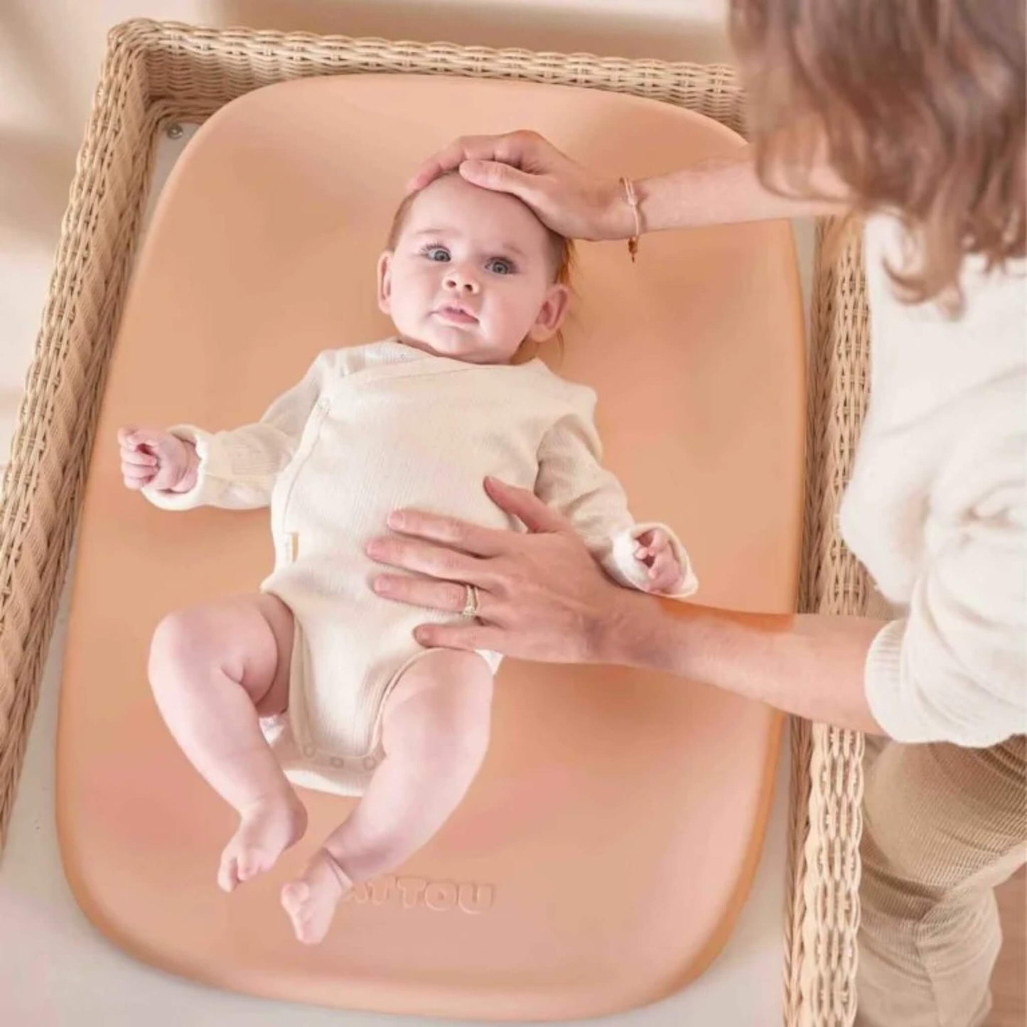 A baby lying on a peach coloured PU foam changing pad inside a woven changing basket while an adult gently supports their head and torso during a nappy change.