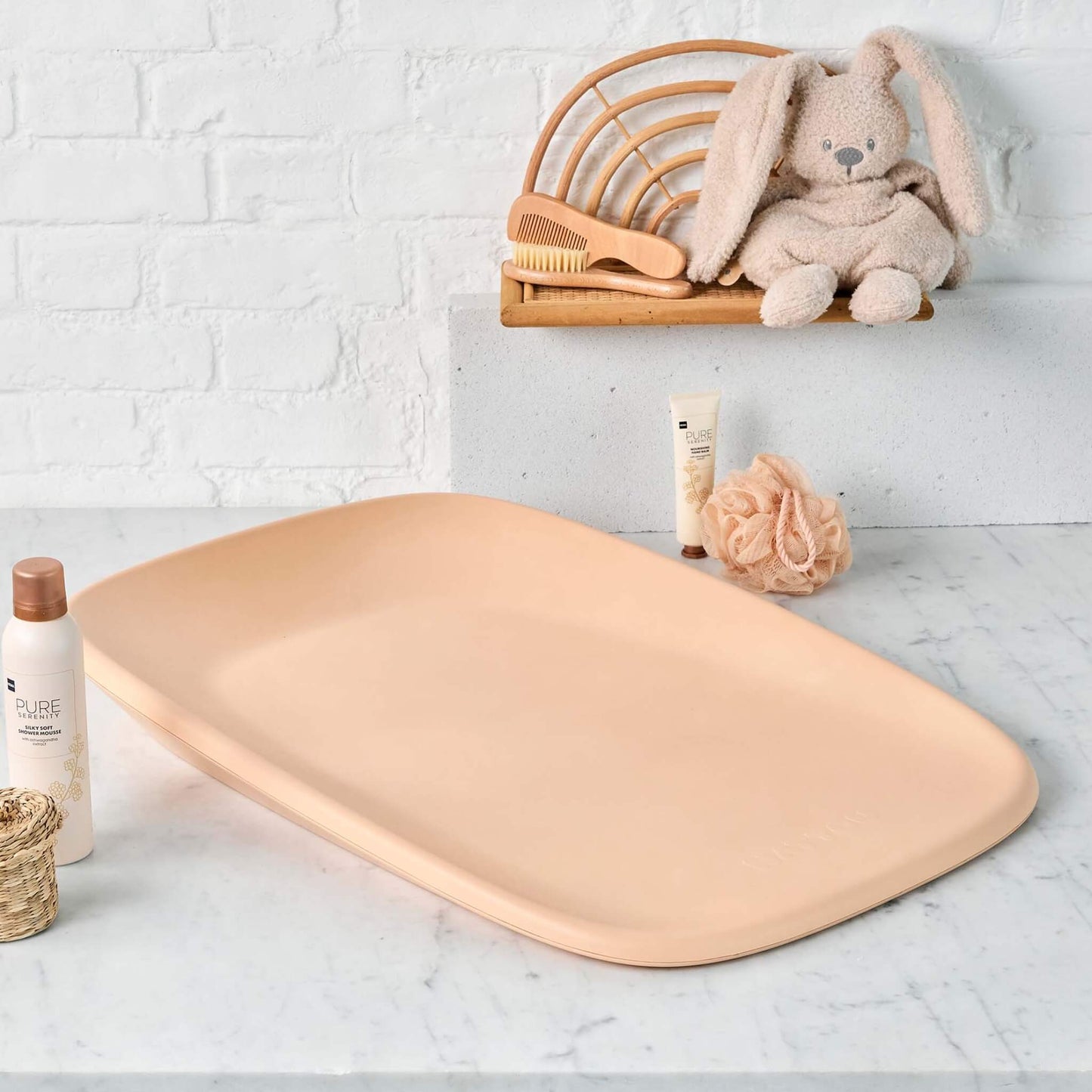A peach baby changing pad placed on a marble countertop, surrounded by baby care items including a brush, lotion, and a plush bunny toy.
