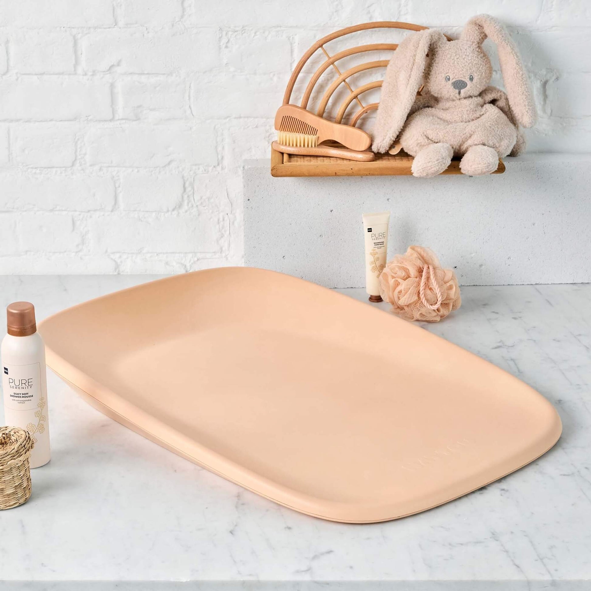 A peach baby changing pad placed on a marble countertop, surrounded by baby care items including a brush, lotion, and a plush bunny toy.