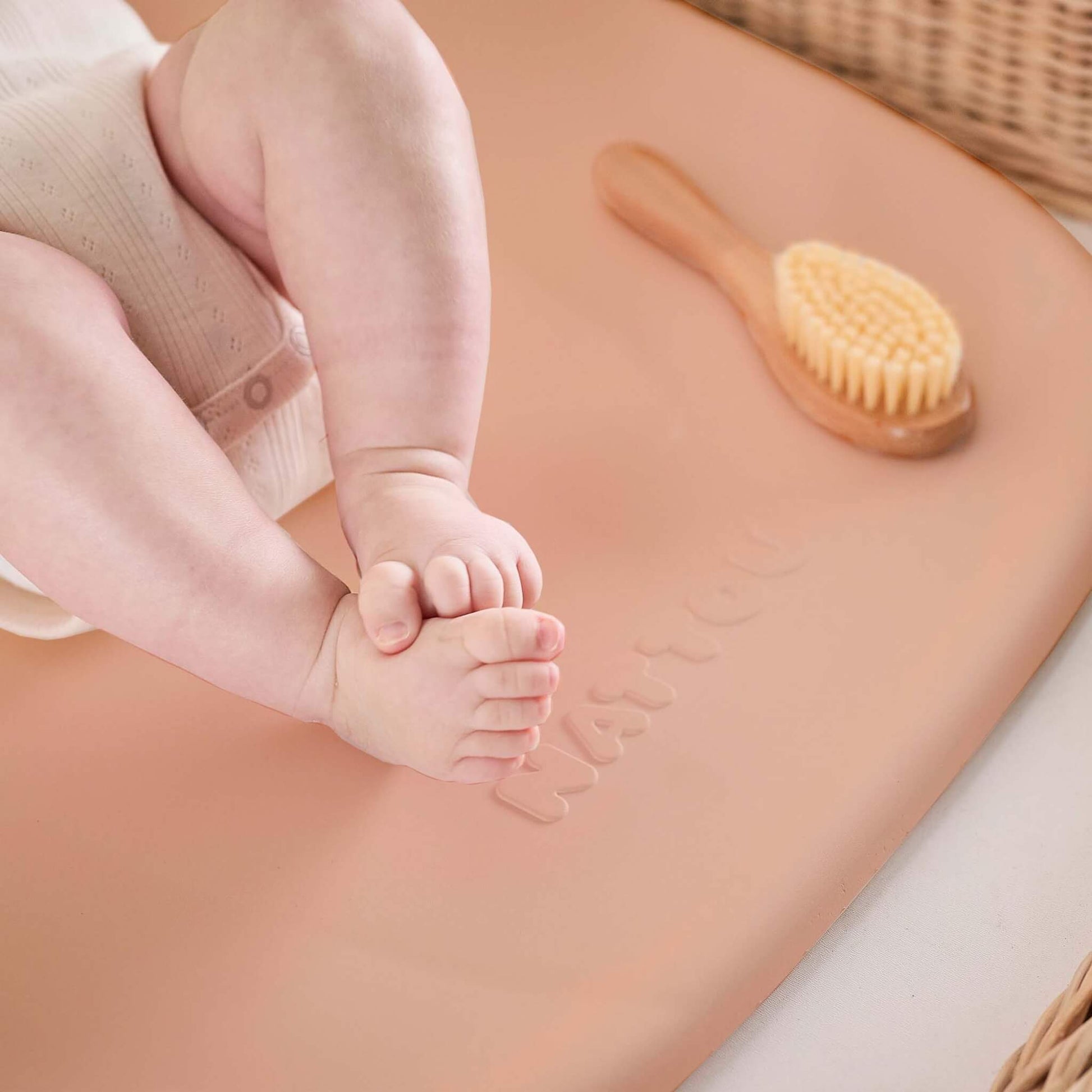 A close-up of a baby’s crossed feet resting on the peach changing pad, with a wooden baby brush placed nearby.