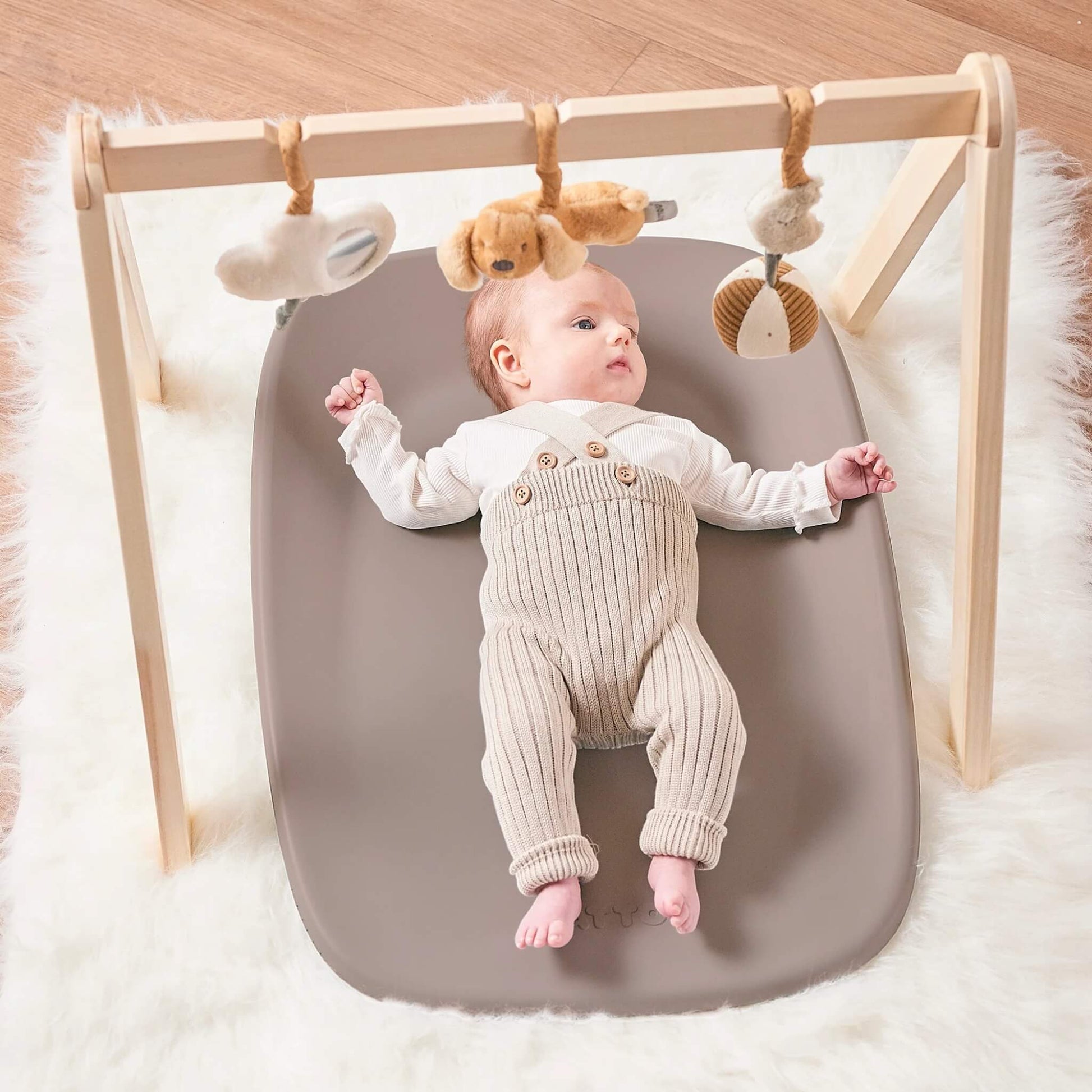Baby lying on a taupe changing pad beneath a wooden play gym with soft hanging toys.