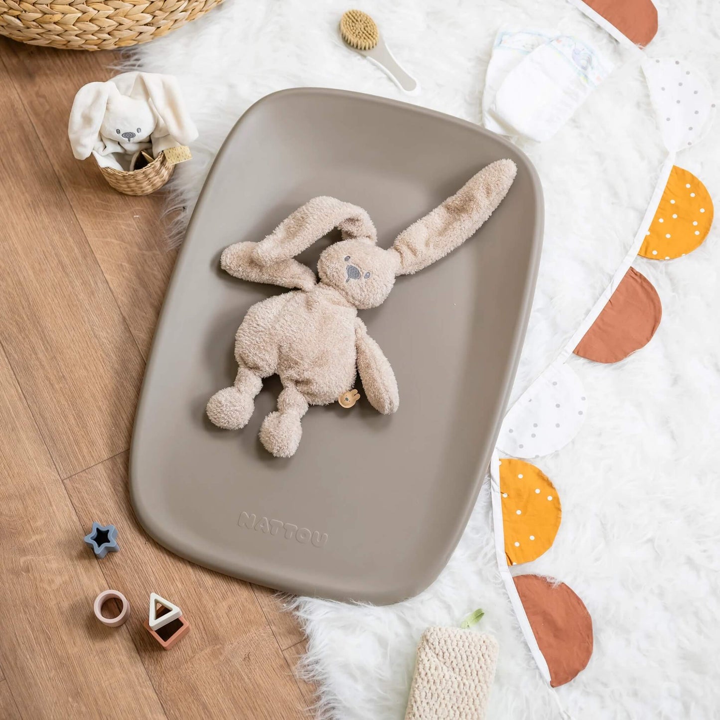 Taupe changing pad placed on a floor with a plush rabbit toy resting on its surface, surrounded by baby accessories.