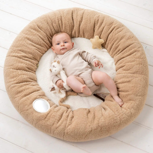 A baby lying inside a round padded playmat with a soft cream centre and a small plush giraffe toy resting beside them.