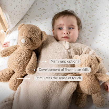 Baby lying in a cot holding a beige teddy bear with another teddy bear on the other side, shown with the text ‘Many grip options’, ‘Development of fine motor skills’ and ‘Stimulates the sense of touch’ on the image.
