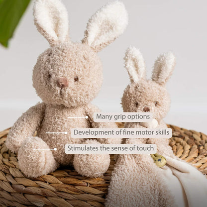 A plush bunny sitting beside a matching comforter on a woven basket, with labels highlighting grip options, fine motor development and tactile stimulation.