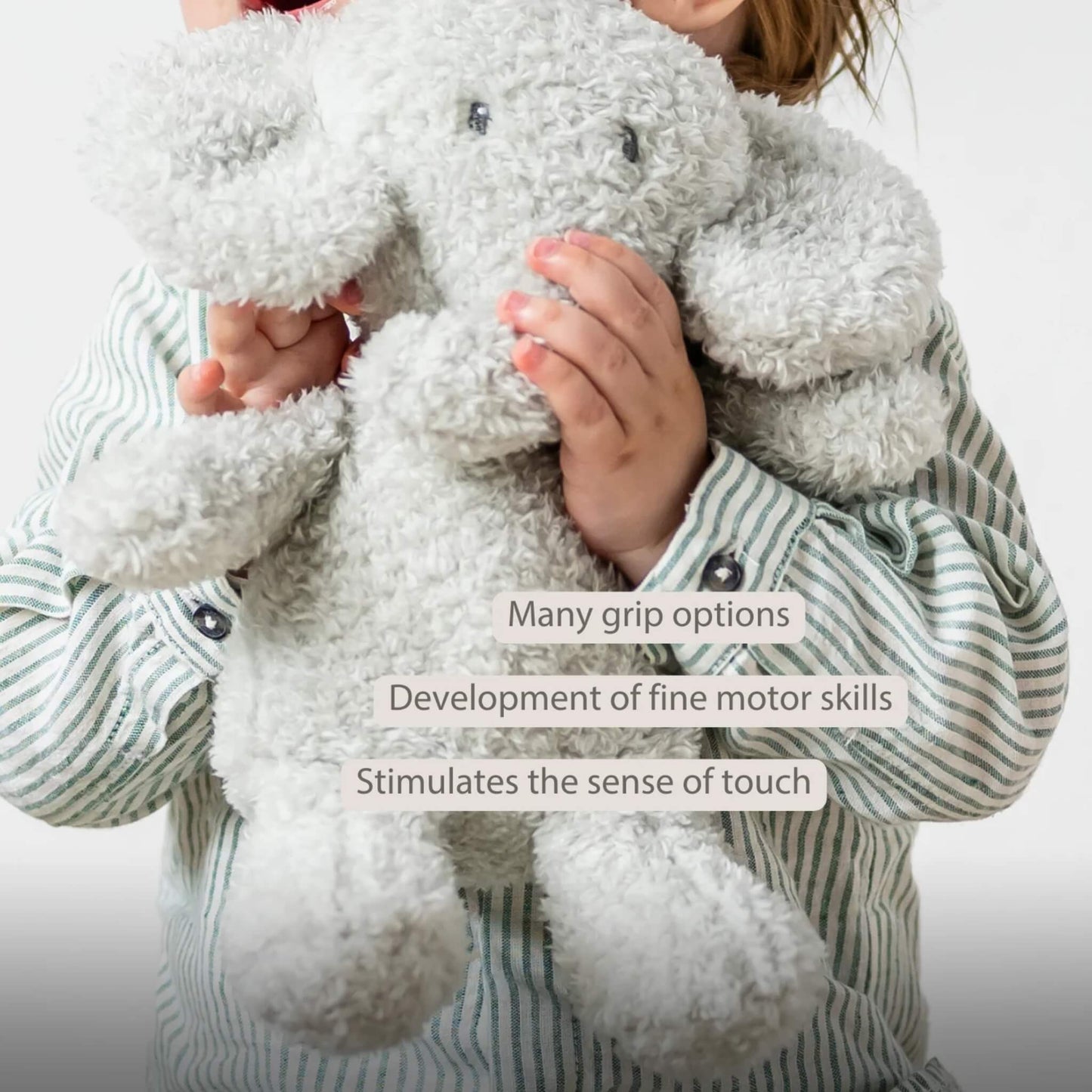 A child holding a plush elephant toy with both hands, showing the toy’s floppy ears and textured teddy fabric. The words “Many grip options”, “Development of fine motor skills” and “Stimulates the sense of touch” appear on the image.