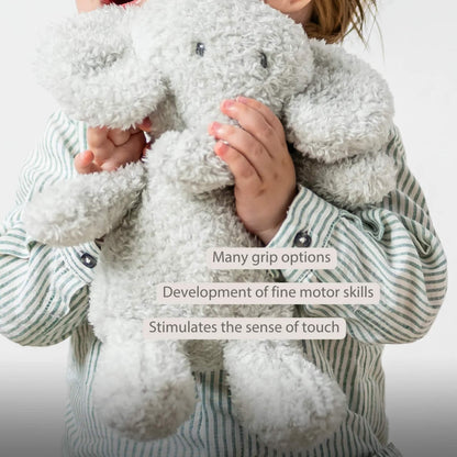 A child holding a plush elephant toy with both hands, showing the toy’s floppy ears and textured teddy fabric. The words “Many grip options”, “Development of fine motor skills” and “Stimulates the sense of touch” appear on the image.