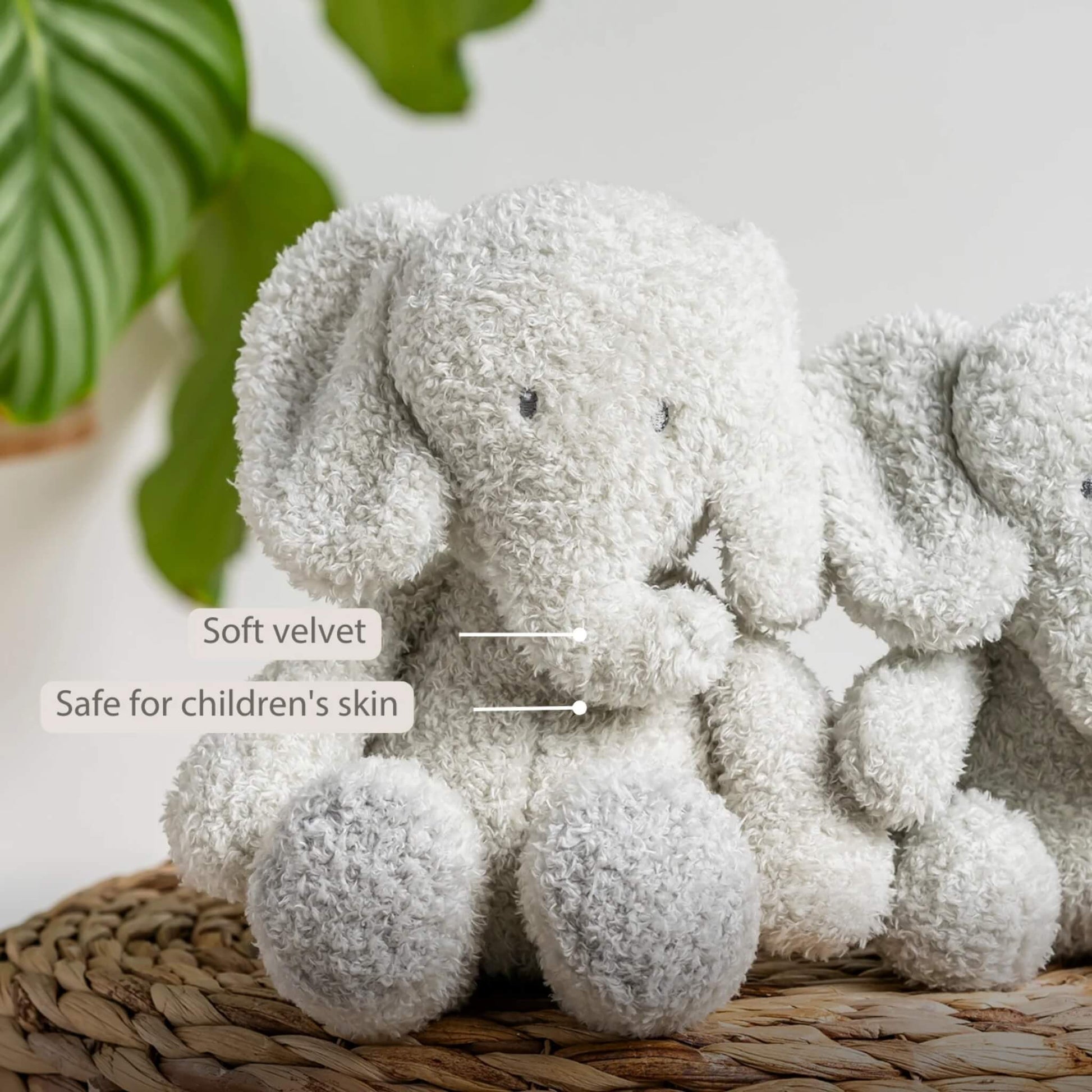 A plush elephant toy placed upright on a woven basket, showing soft rounded features and a fluffy grey finish. The words “Soft velvet” and “Safe for children’s skin” appear on the image.