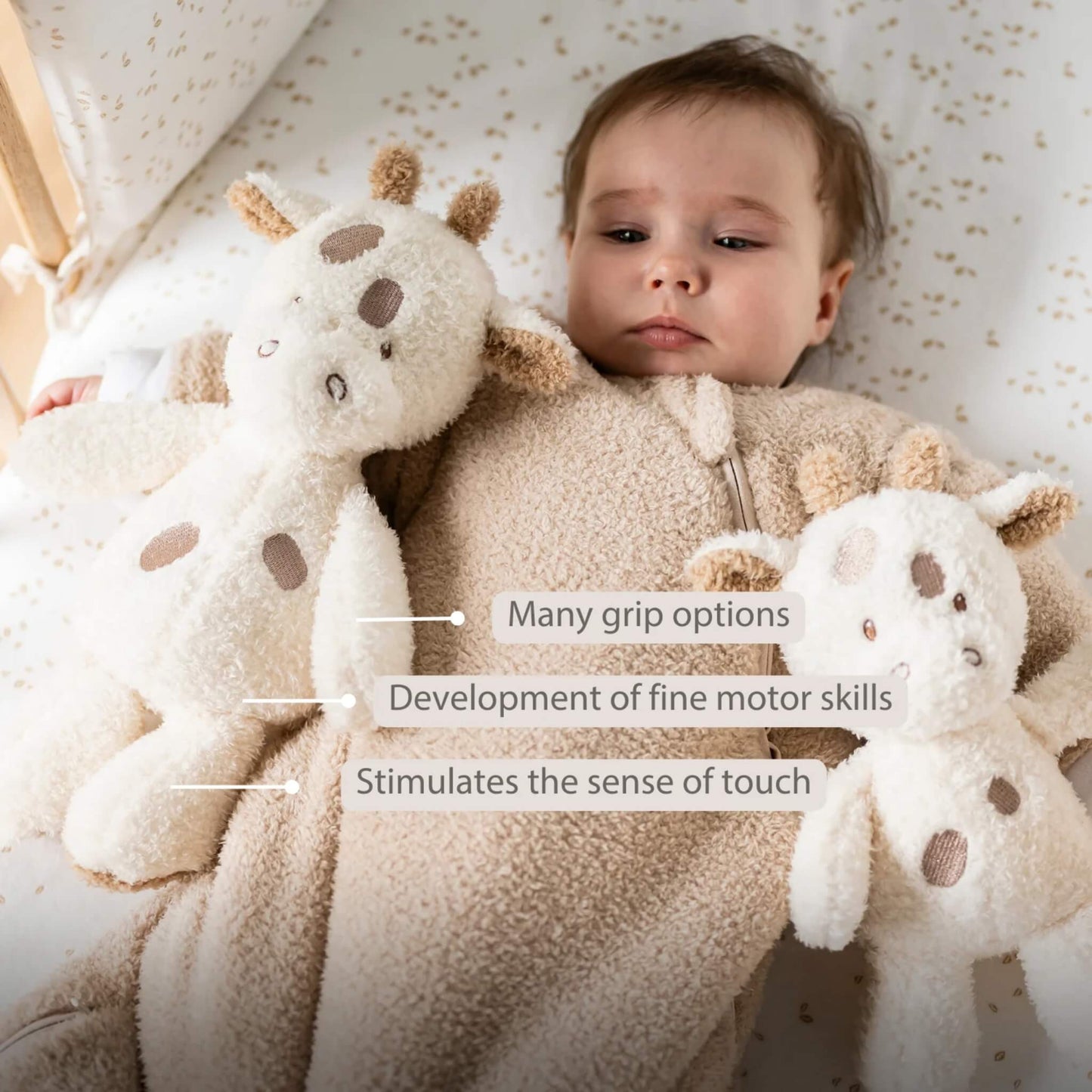 A baby lying on a patterned sheet holding a soft giraffe plush toy in each hand, with on-image text reading “Many grip options”, “Development of fine motor skills” and “Stimulates the sense of touch.” The toys have cream fur, stitched features and brown embroidered spots.