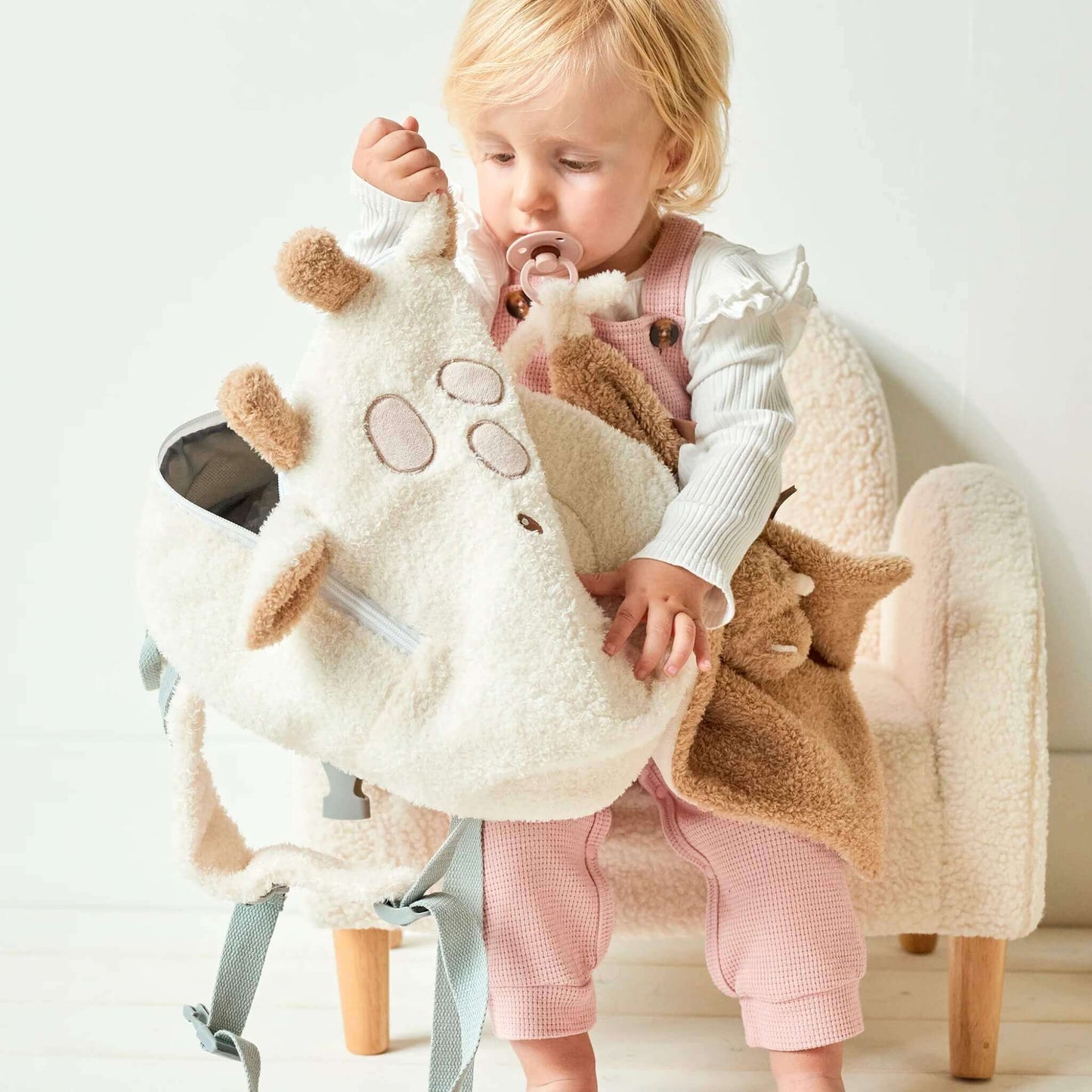 A toddler sitting on a small chair while holding an open giraffe-shaped backpack, revealing the soft fabric and spacious interior.