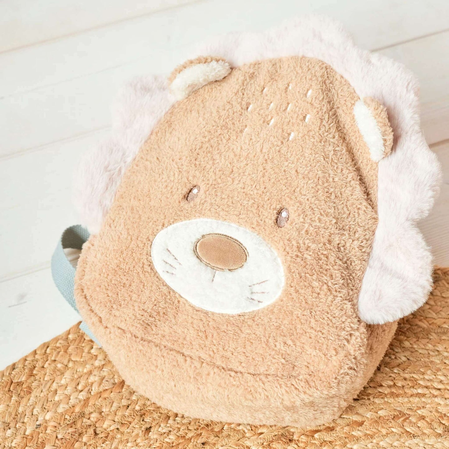A lion-shaped toddler backpack resting on a woven mat, showing its soft textured fabric, plush mane and small embroidered features.