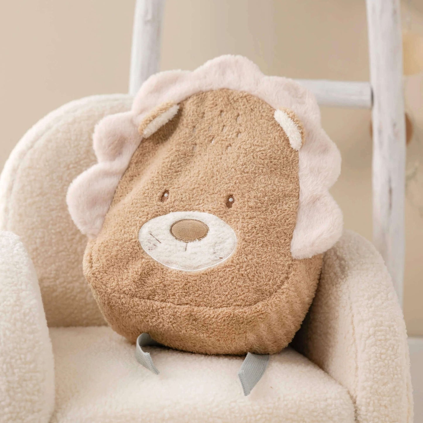 A lion-shaped toddler backpack placed on a cream textured chair, showing the plush mane, embroidered face and adjustable grey straps.