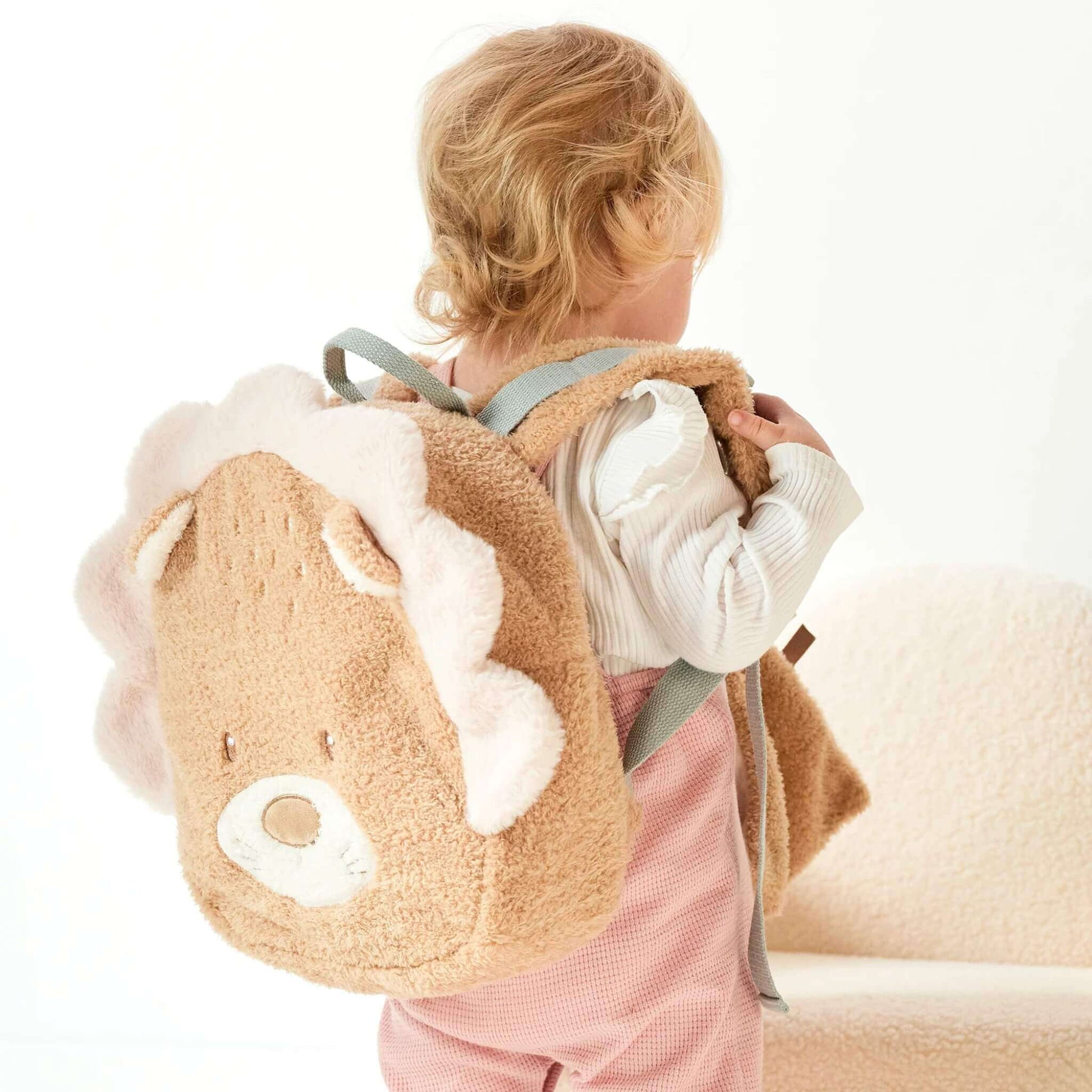 A toddler wearing a lion-shaped backpack with plush mane and grey straps, seen from behind while standing in a bright room.