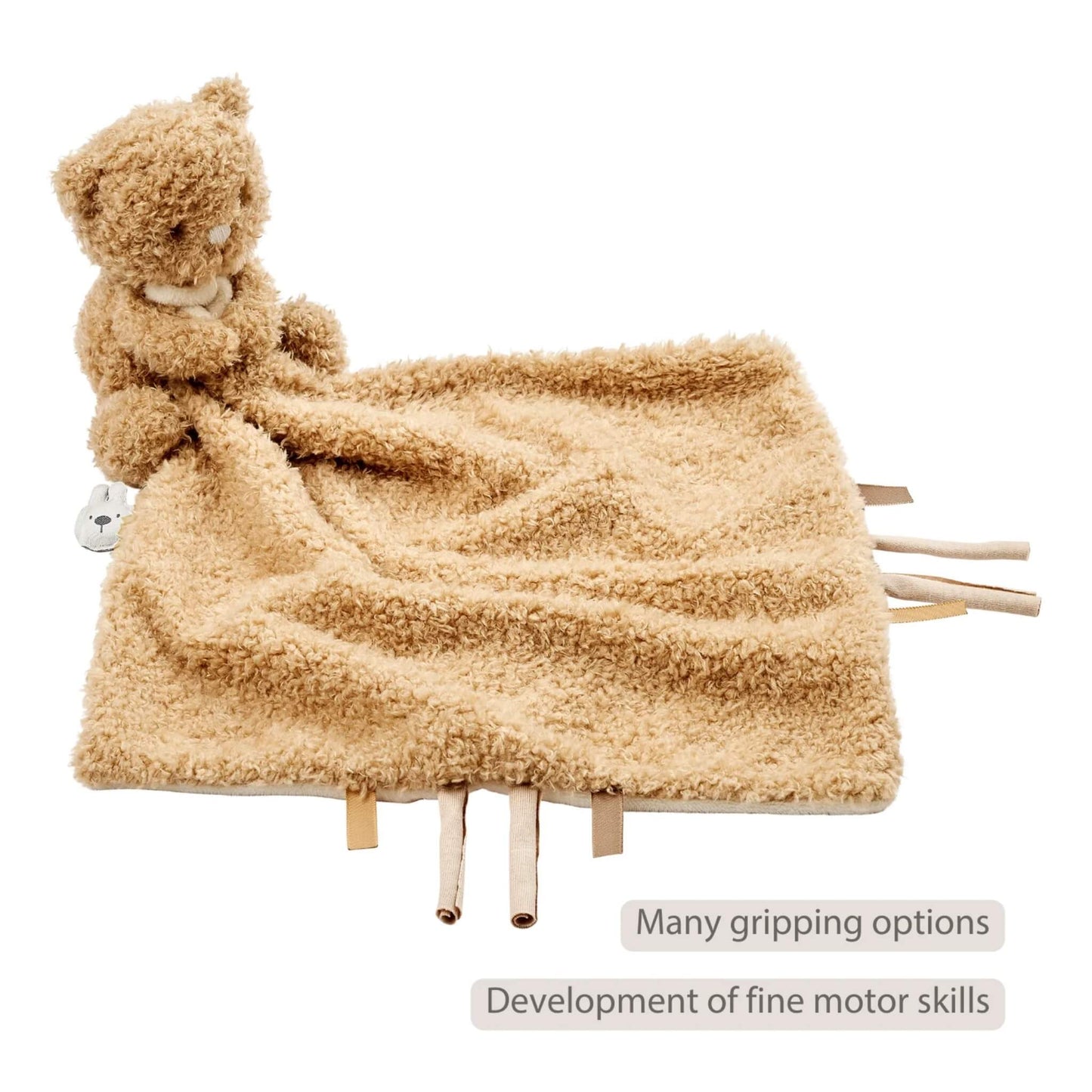 Cuddly beige baby comforter with an attached plush bear shown with fabric tags along the edges, displayed with the text ‘Many gripping options’ and ‘Development of fine motor skills’ on the image.