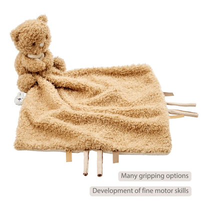 Cuddly beige baby comforter with an attached plush bear shown with fabric tags along the edges, displayed with the text ‘Many gripping options’ and ‘Development of fine motor skills’ on the image.