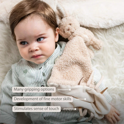 Baby lying on a fluffy surface while holding a beige cuddle cloth with fabric tags, shown with the text ‘Many gripping options’, ‘Development of fine motor skills’, and ‘Stimulates sense of touch’ on the image.