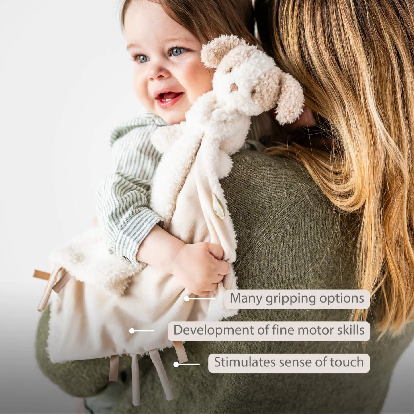 Baby holding a beige security with an attached plush dog while being carried, shown with the text ‘Many gripping options’, ‘Development of fine motor skills’ and ‘Stimulates sense of touch’ on the image.